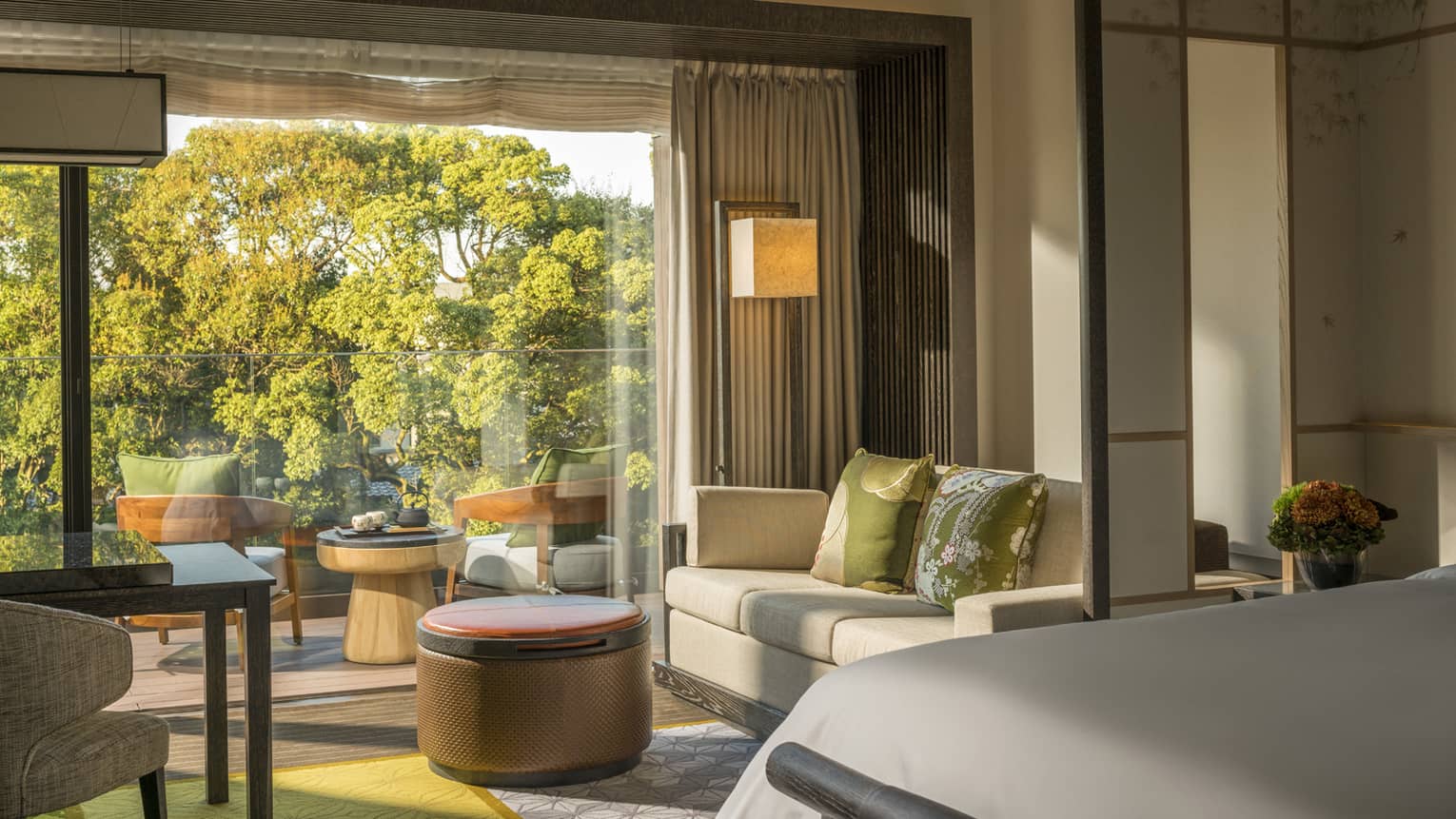 Guest room with king bed, sofa, desk and chair, round ottoman and private balcony with a seating area overlooking a pond garden
