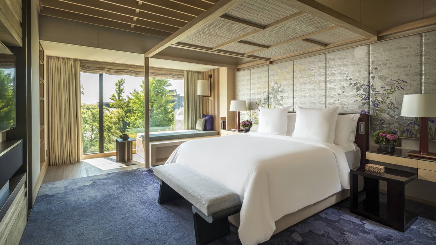 Luxury bedroom with a king bed, tufted bench and floor-to-ceiling windows overlooking lush greenery