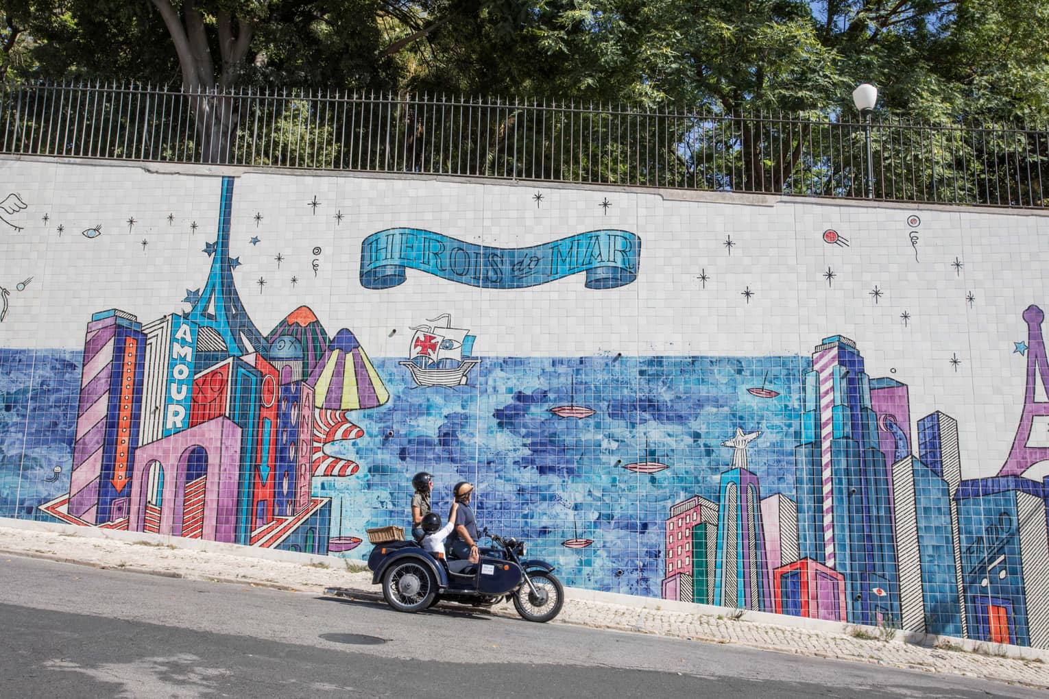 Two adults and a child stop their motorcycle to look at a city mural made of tiles