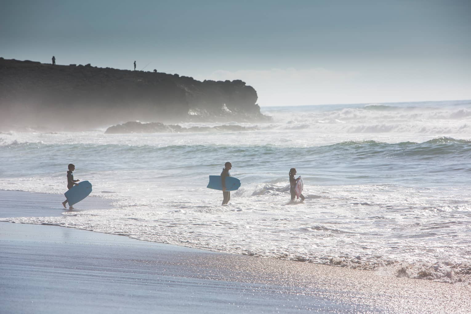 Silhouettes of man, woman and child all wearing wetsuits, holding blue surfboards, walking into ocean
