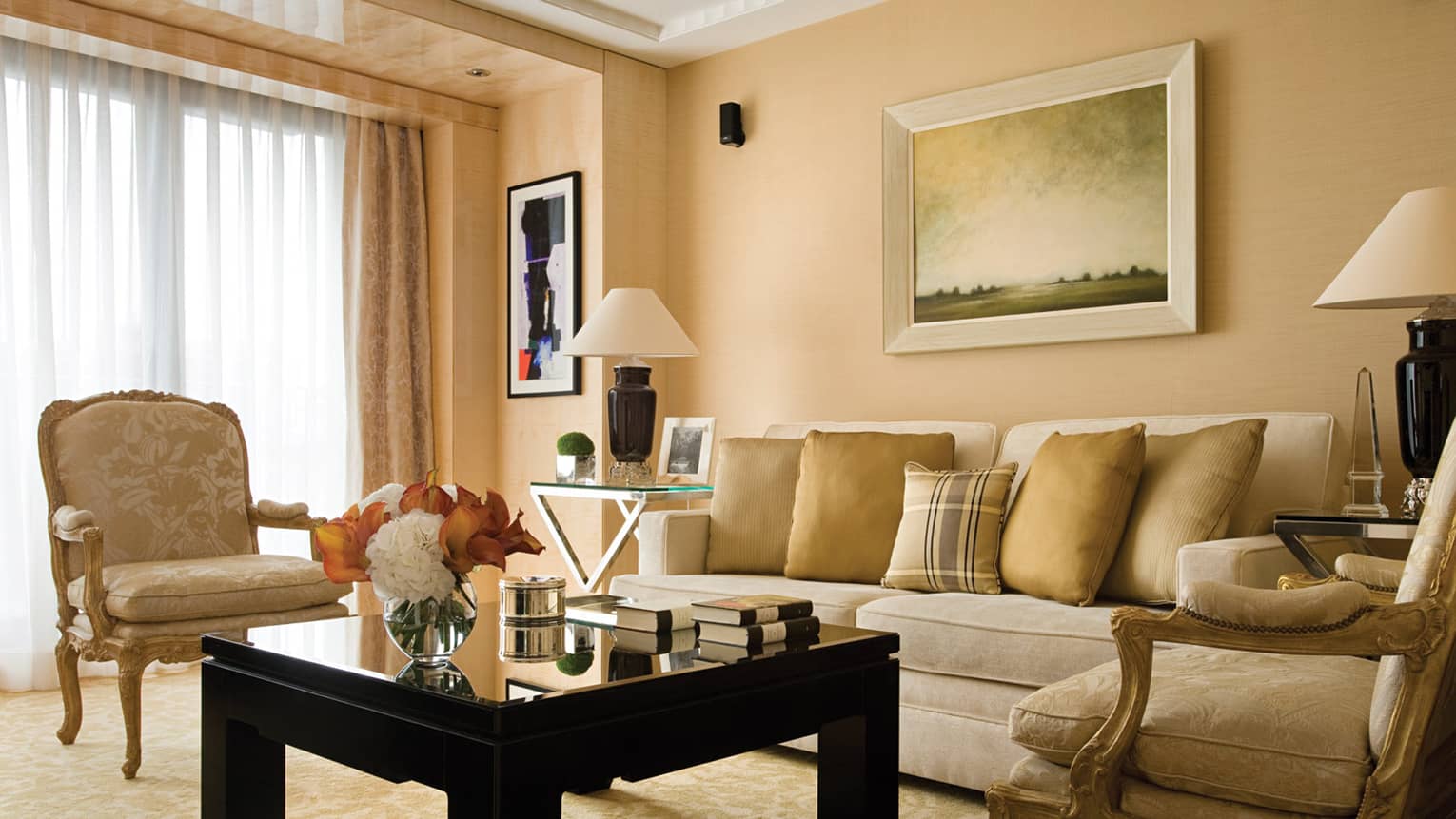 One-bedroom Suite living room with gold accent chairs, flowers and books on black coffee table,One-bedroom Suite living room with gold accent chairs, flowers and books on black coffee tableThe Deluxe Suite at Four Seasons London Park Lane in Mayfair showcases a classic and luxurious living room setting. The room features a palette of soft beige and gold tones, with a plush beige sofa adorned with gold cushions. Two ornate, antique-style armchairs flank a sleek black coffee table, creating a sophisticated and inviting conversation area. Large windows are draped with heavy curtains, allowing natural light to enhance the warm atmosphere. The decor is completed by a traditional landscape painting above the sofa and elegant table lamps.