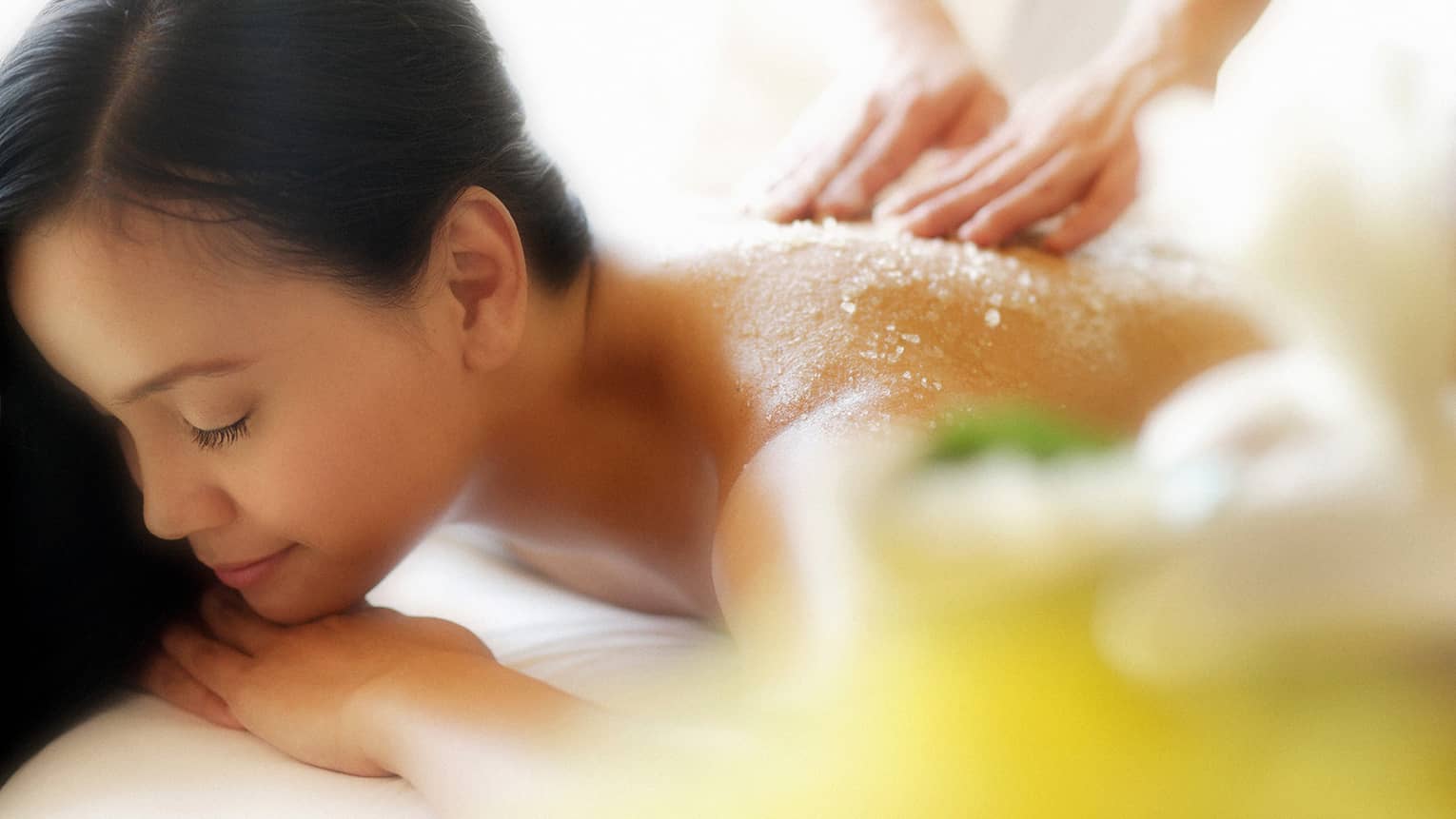 Woman lying down with eyes closed as hands massage salt scrub into shoulders