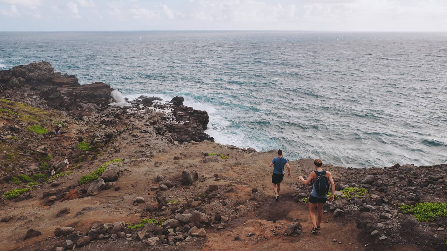 Two people hike down dirt-covered path, rocks towards ocean