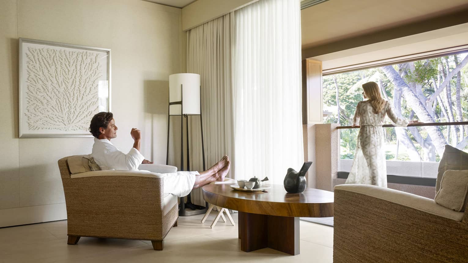Man in white bathrobe kicks back in armchair in Spa room, woman in sheer robe stands at sunny window