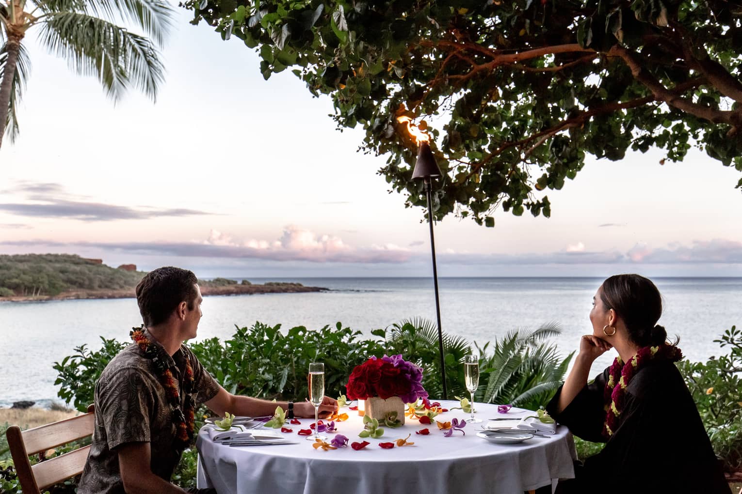 A couple wearing leis at a white table, and looking out on the ocean.