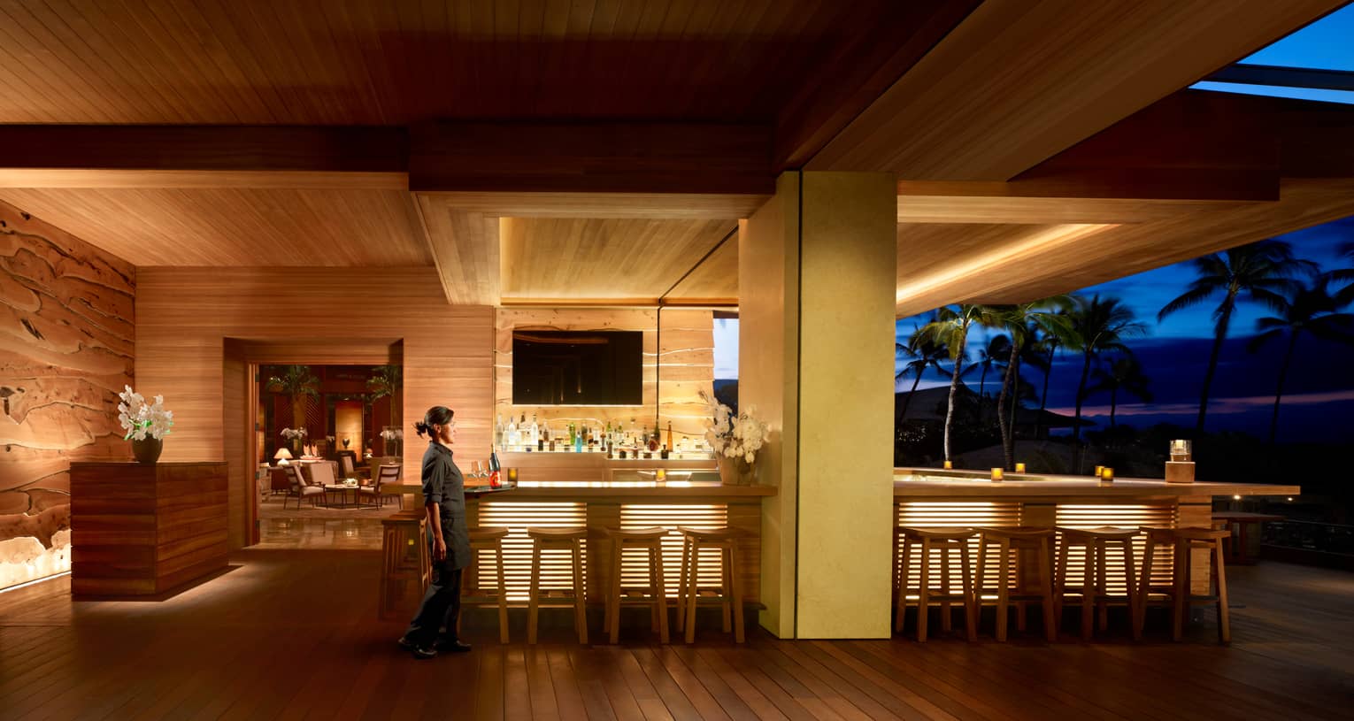 Server walks by wood NOBU Lanai restaurant bar with lights on indoor outdoor patio at night
