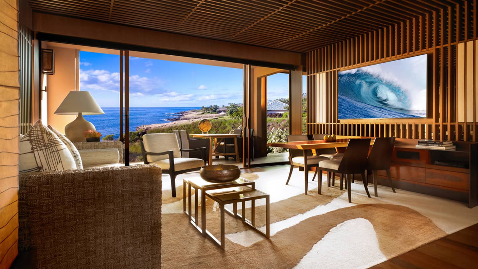 Ocean view suite open concept with tiki-style rattan furniture, white cushions, white-and-brown area rug, glass wall to balcony