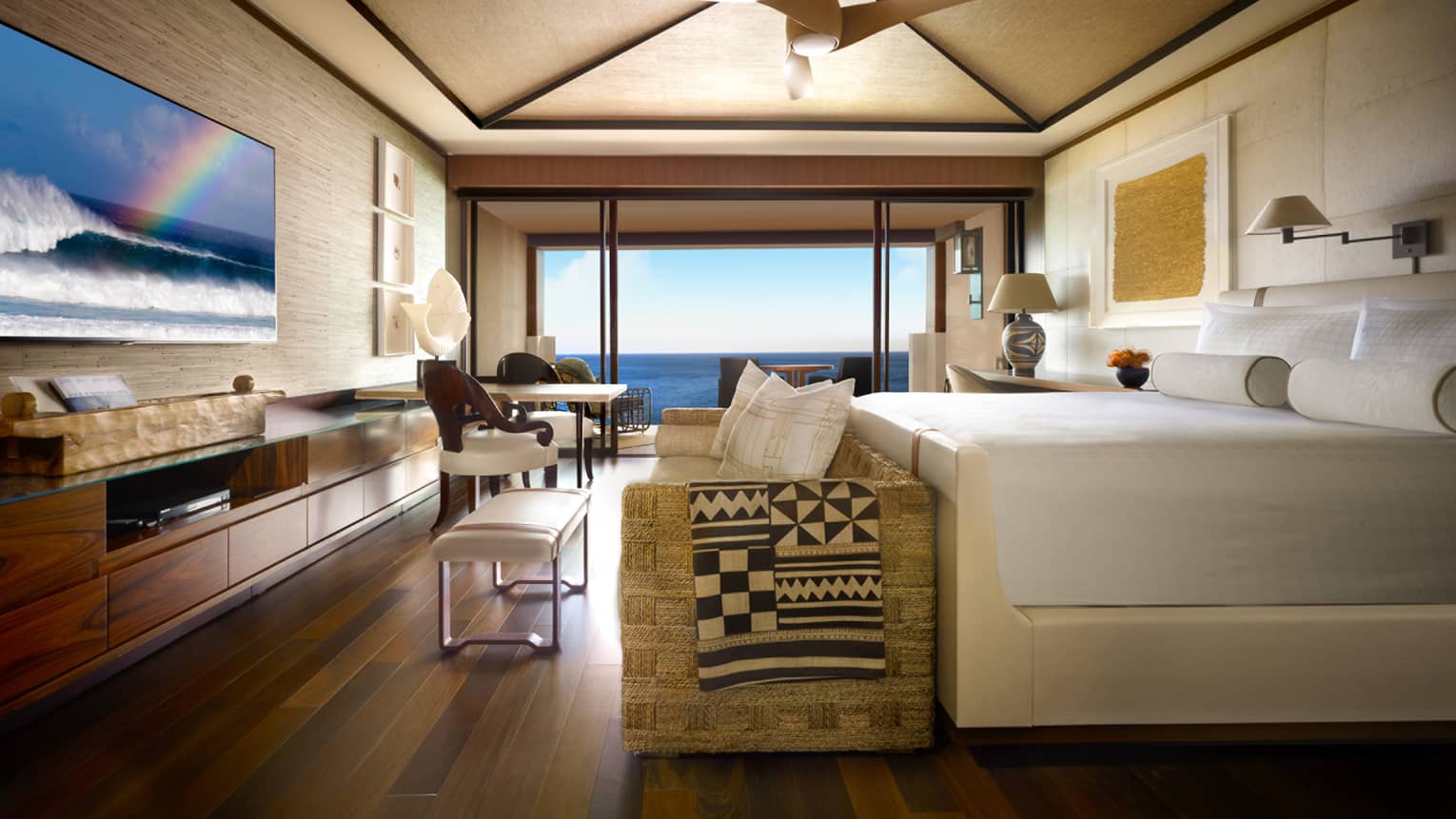 Prime oceanfront room bed with white frame, rattan sofa, long wood dresser, flat-screen tv with rainbow, glass balcony window