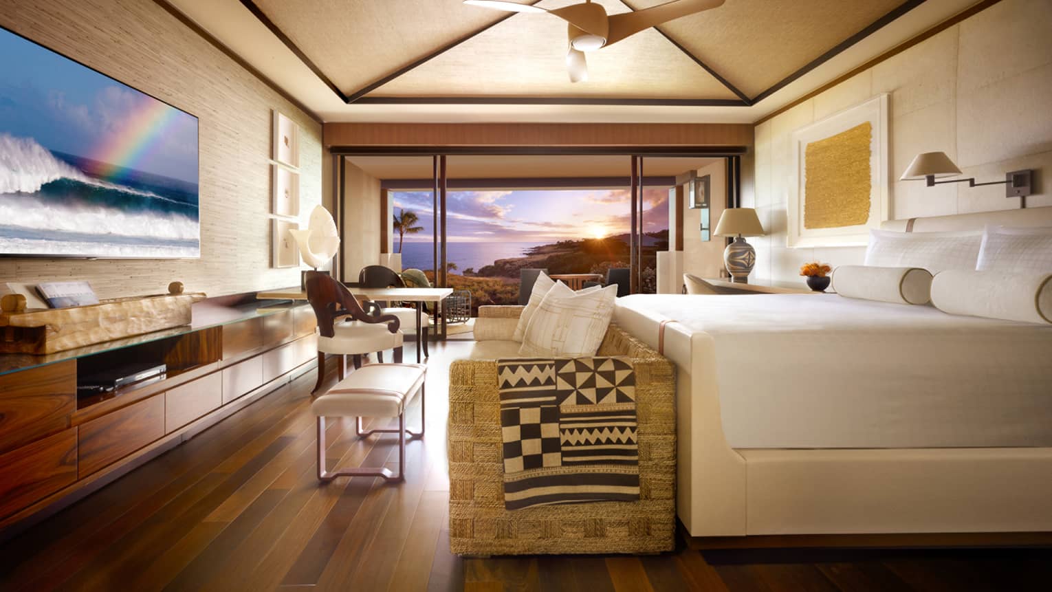Partial ocean-view room with rattan sofa and folded blanket in front of bed, large flat-screen TV with rainbow, balcony window