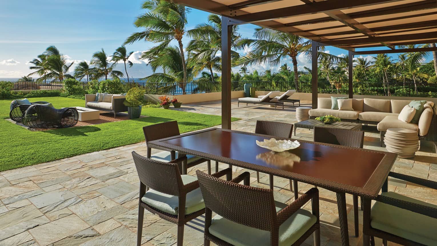 Outdoor patio with dining table, cushioned lounge seating and sun loungers, overlooking a lush garden and palm trees at a tropical resort