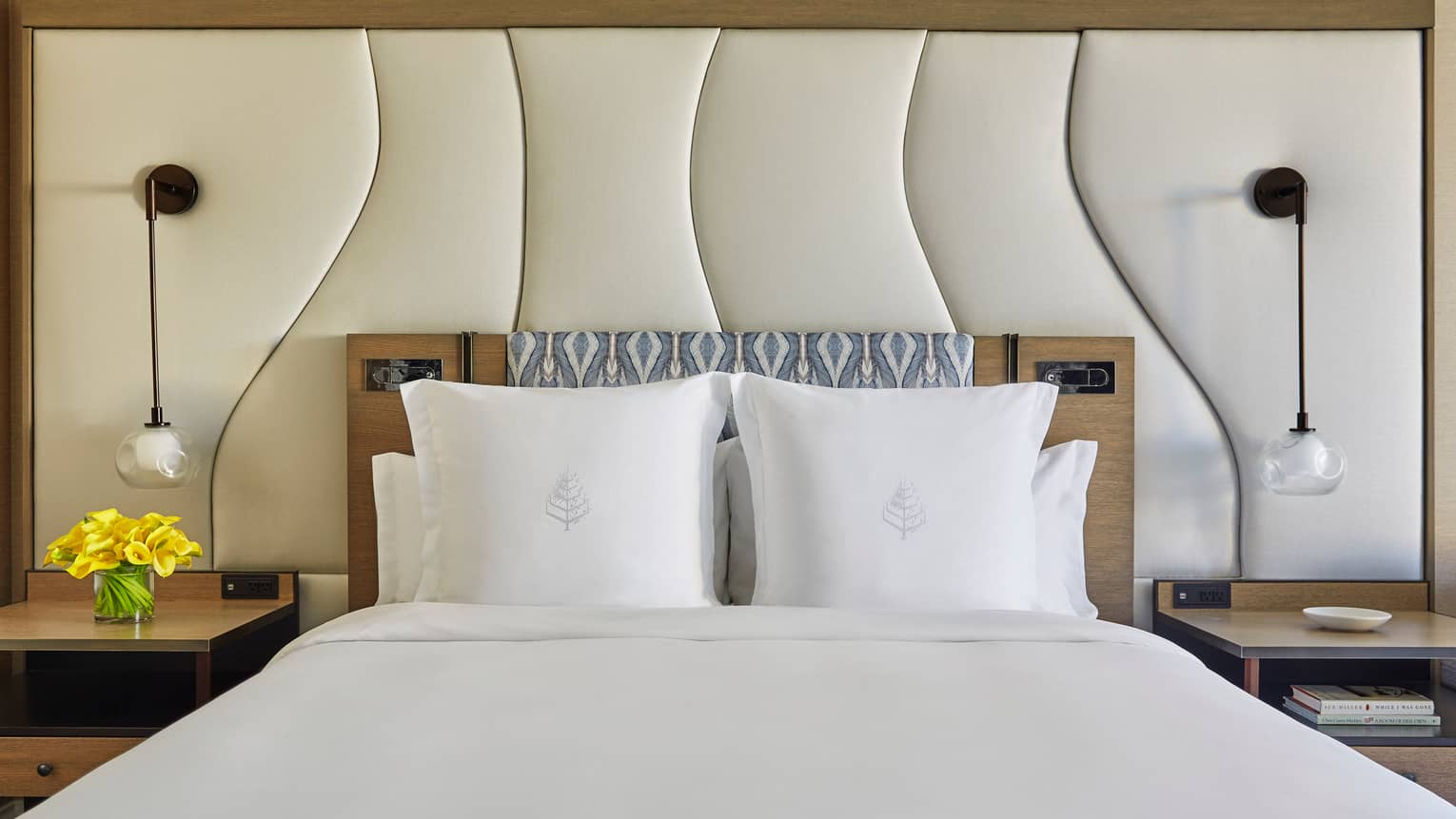Four Seasons bed with white linens, padded headboard, pendant lights and bedside tables with yellow flowers on the left