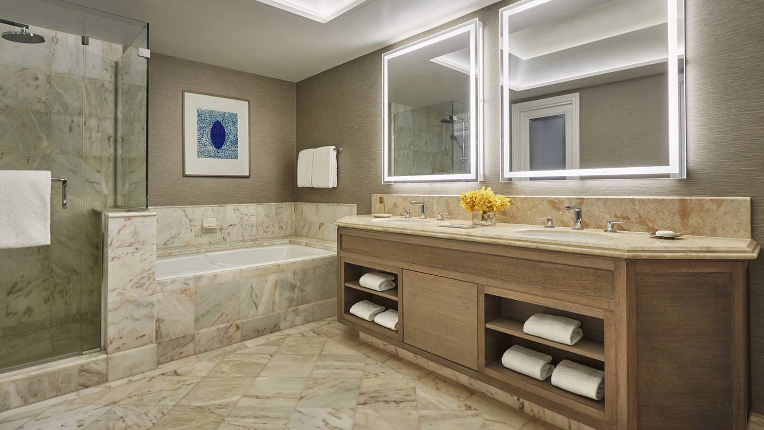 Luxury bathroom with marble floors, bathtub, glass-enclosed shower, double vanity with illuminated mirrors and yellow flowers
