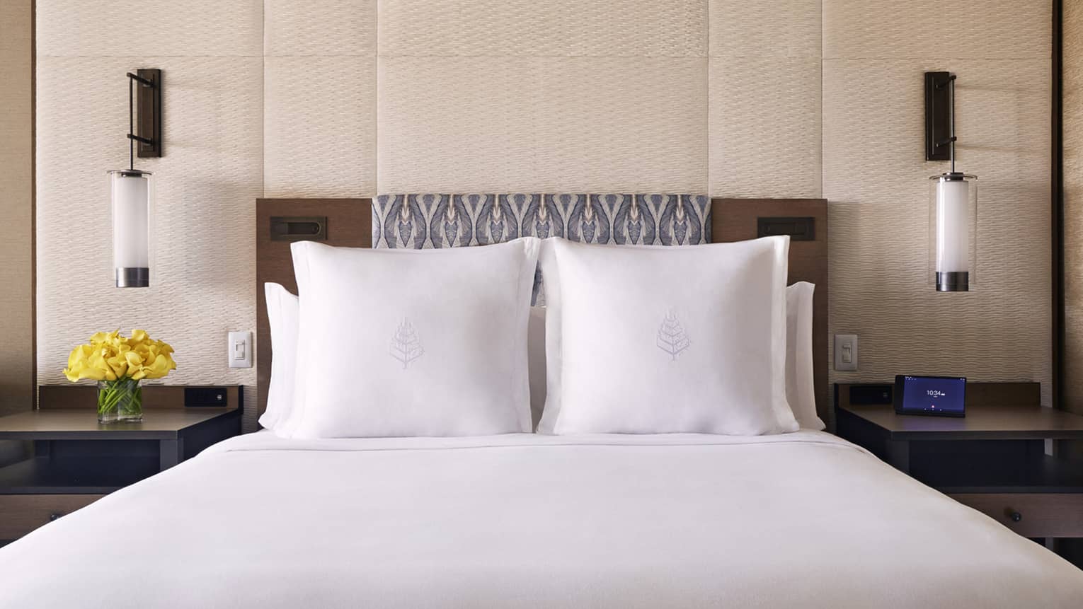 Four Seasons bed with white linens, two pillows, bedside tables with yellow flowers and electronic device and modern pendant lights