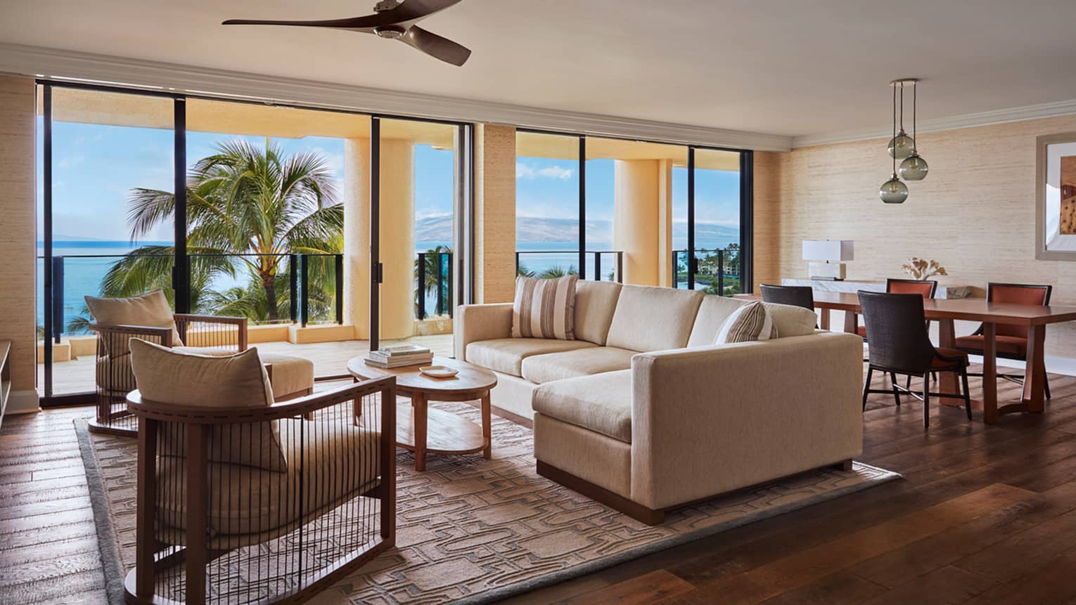 Spacious living and dining area with a beige sofa, armchairs and dining table, opening to a balcony with ocean and palm trees view at a luxury resort suite