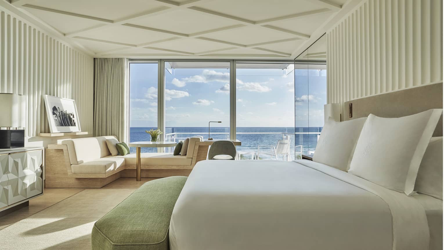 Luxury hotel room with a king bed, light-coloured furnishings and floor-to-ceiling windows offering a view of the ocean and blue sky