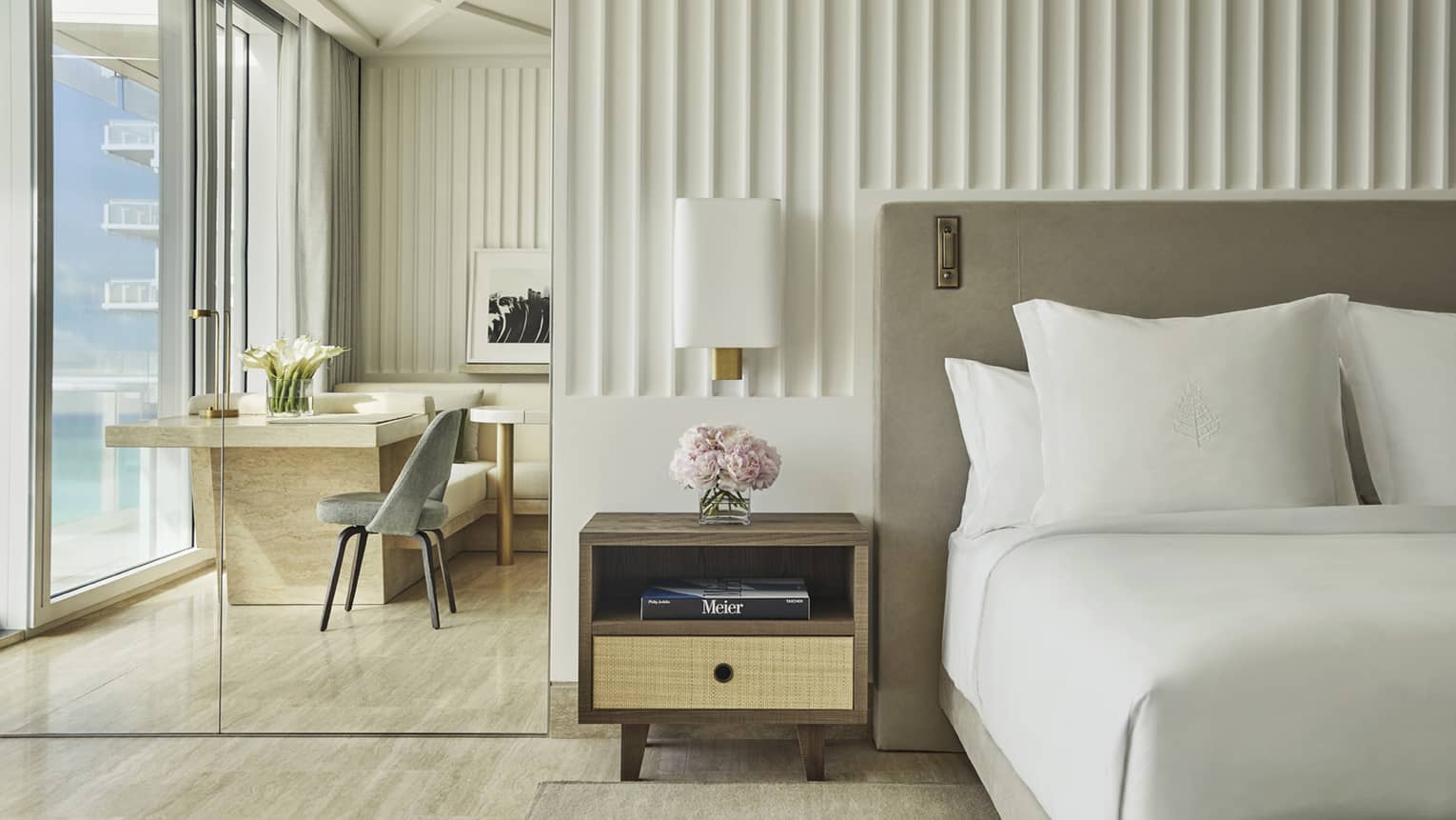 Elegant hotel room with a neatly made bed, a bedside table with flowers and a desk area by floor-to-ceiling windows