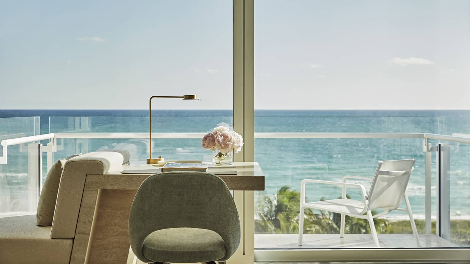 Luxury oceanfront hotel room desk with a chair, a lamp and pink flowers, overlooking a balcony with ocean views under a clear sky
