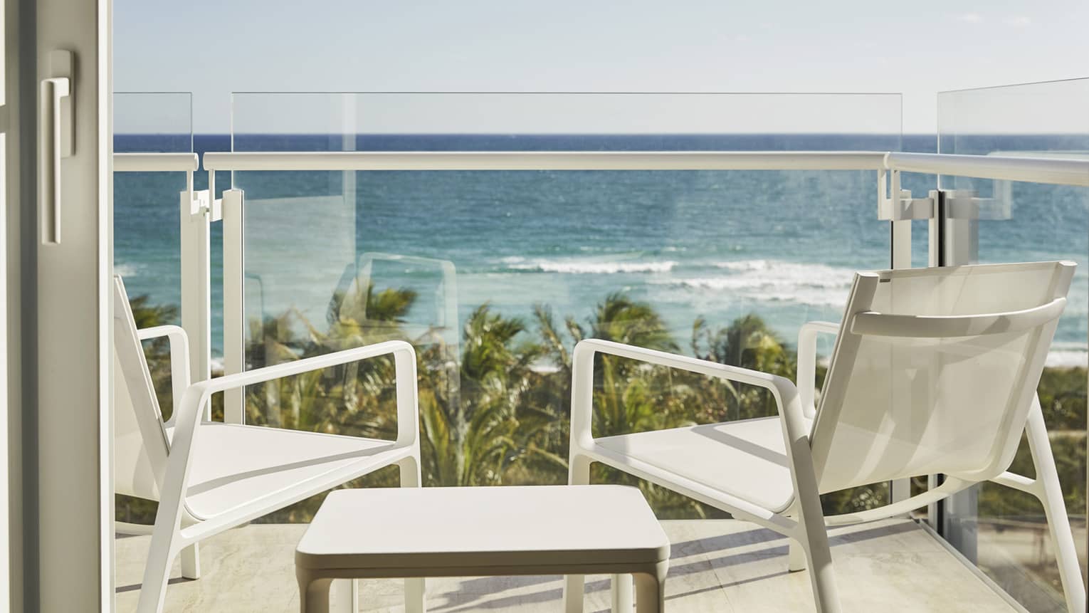 Luxury hotel balcony with two white chairs and a table overlooking the ocean and palm trees under a clear blue sky