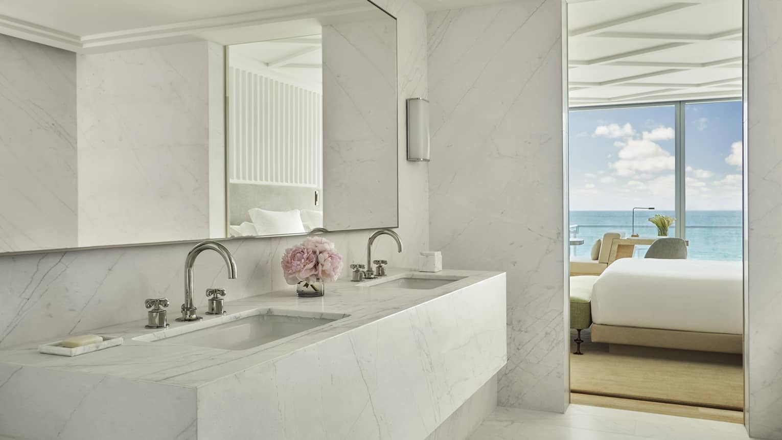 Luxury hotel bathroom with a marble double sink vanity, a mirror and a view into the bedroom with ocean views through floor-to-ceiling windows