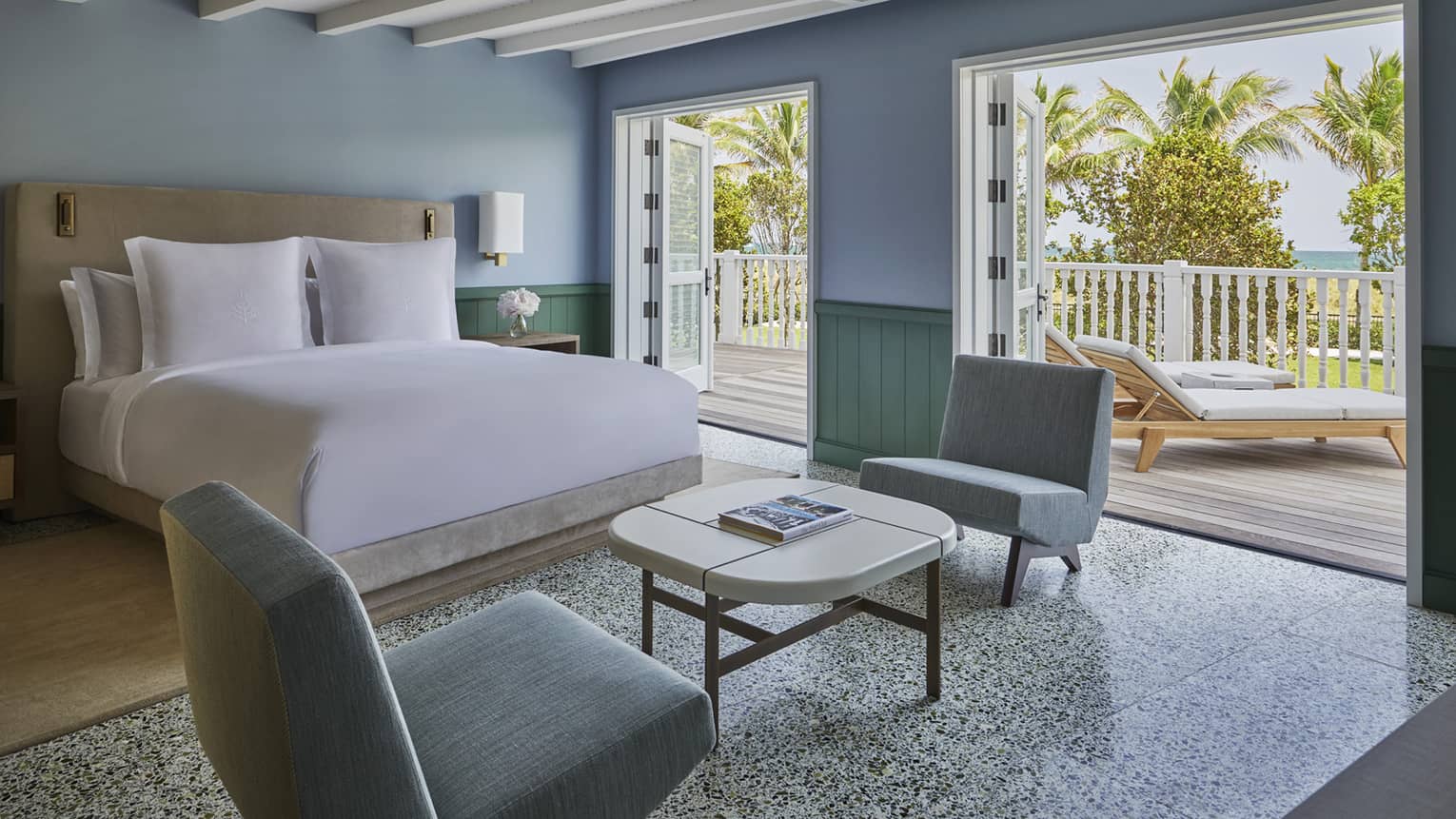 Luxury hotel room with a king bed, blue-toned walls, two armchairs, coffee table and open doors leading to a balcony with lounge chairs and a view of palm trees