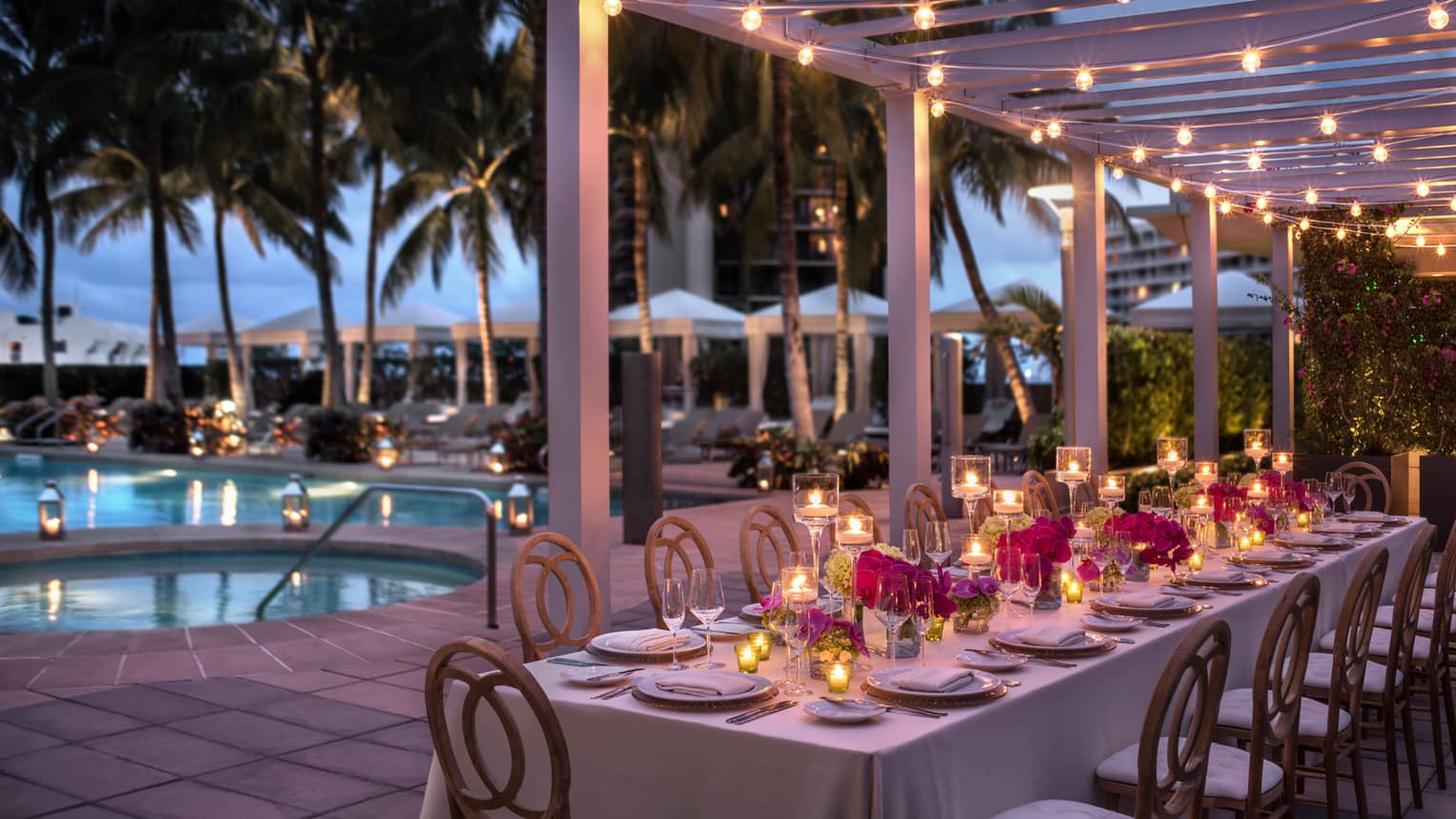 Long, candle-lit dining table with flowers by outdoor pool at dusk