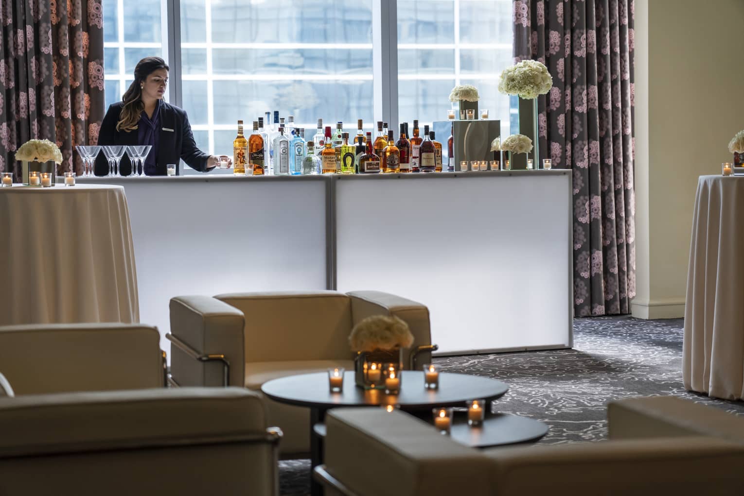 Bartender stands at liquor bottle-lined bar in front of window near seating area