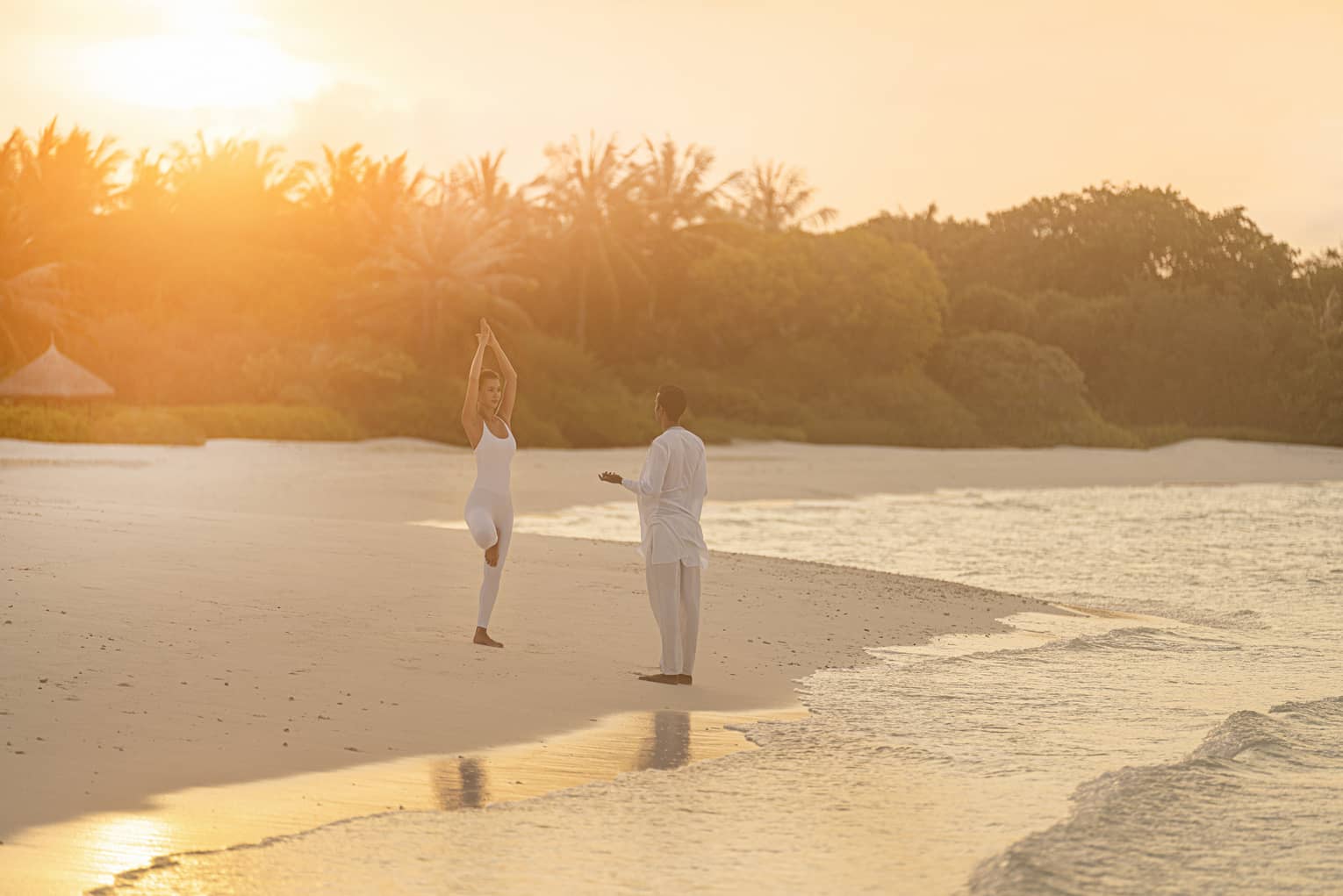 Person practicing yoga on a tropical beach at sunrise with an instructor, surrounded by calm ocean waves and lush greenery in the background