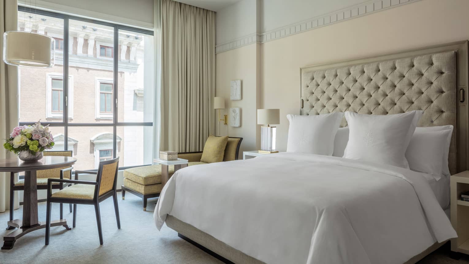 Four Seasons Hotel Madrid's Superior room is elegantly designed with a cream tuffed headboard, white bed linens, a floor to ceiling window with cream curtains and yellow chairs.  