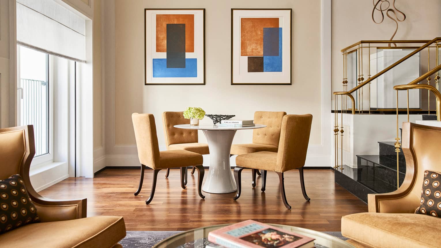 Hotel suite dining area with a round white table, four mustard upholstered chairs, modern wall art, gold-accented staircase and natural light from large windows