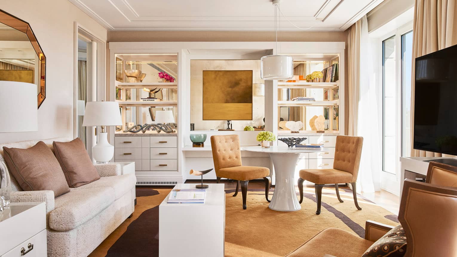Luxury hotel living room with plush seating, tan chairs, round and rectangular tables, TV, decorative shelves and large windows with beige curtains