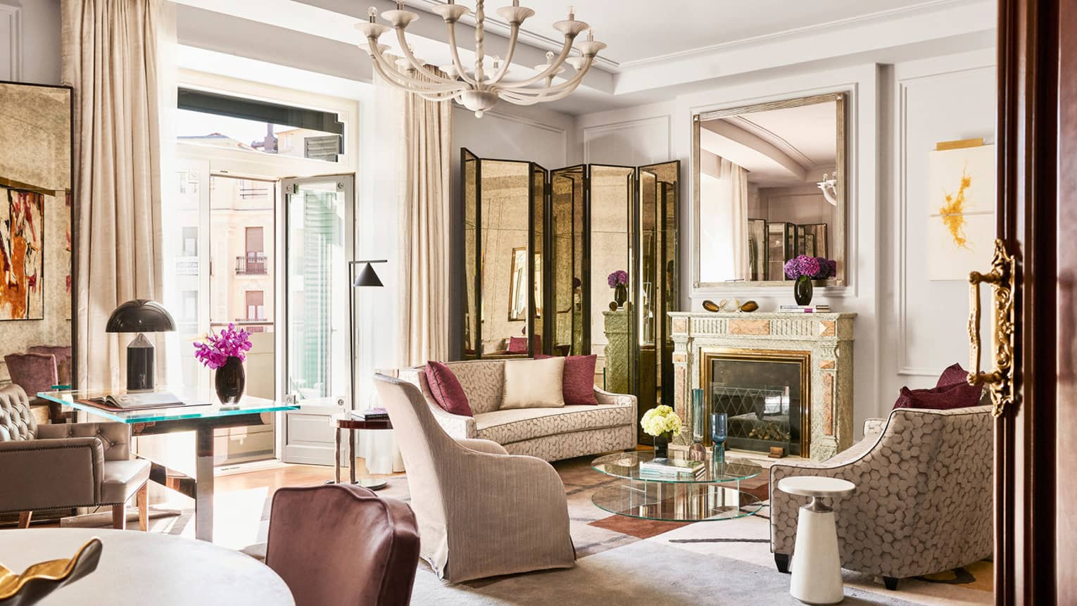 Luxury hotel suite with upholstered seating, glass coffee table, desk, dining table, decorative fireplace, large mirror, chandelier and open doors to balcony