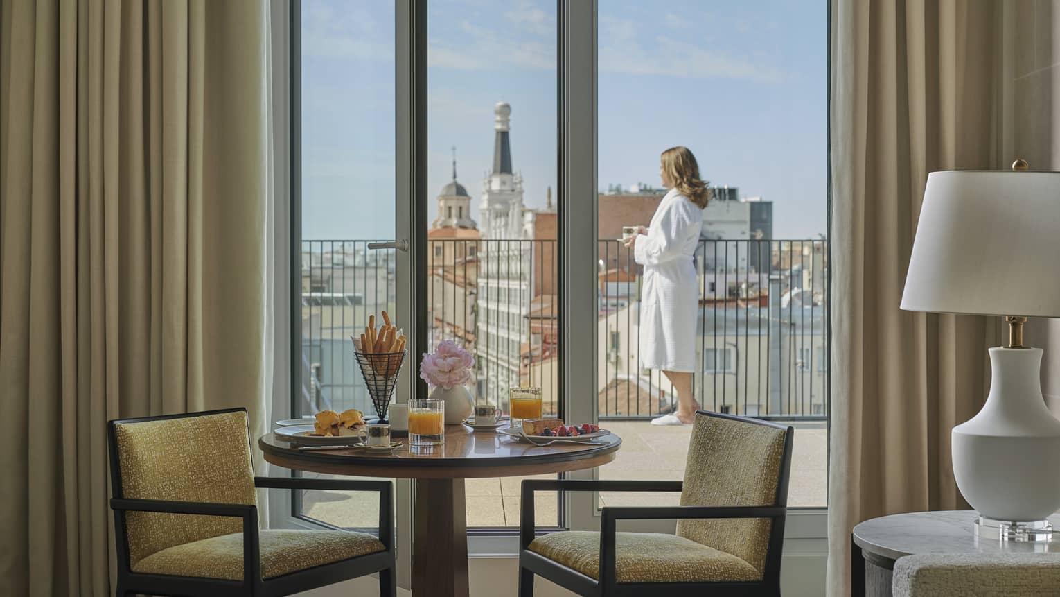 Luxury hotel room breakfast setup with juice, pastries and coffee on a table by balcony doors with a person in a robe enjoying a city view from a terrace