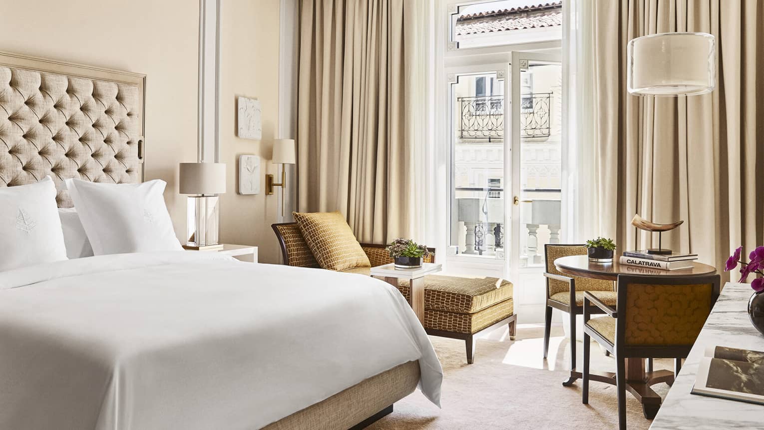 Luxury hotel bedroom with king bed, tufted headboard, table with books and horn sculpture, tan chairs and chaise lounge and doors opening to a Juliet balcony