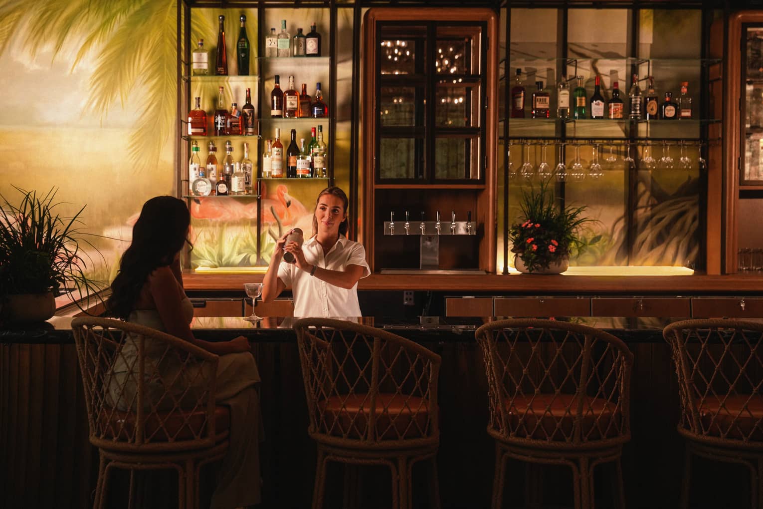 Bartender mixes a drink for a guest at a bar with a mural of a beach on the wall