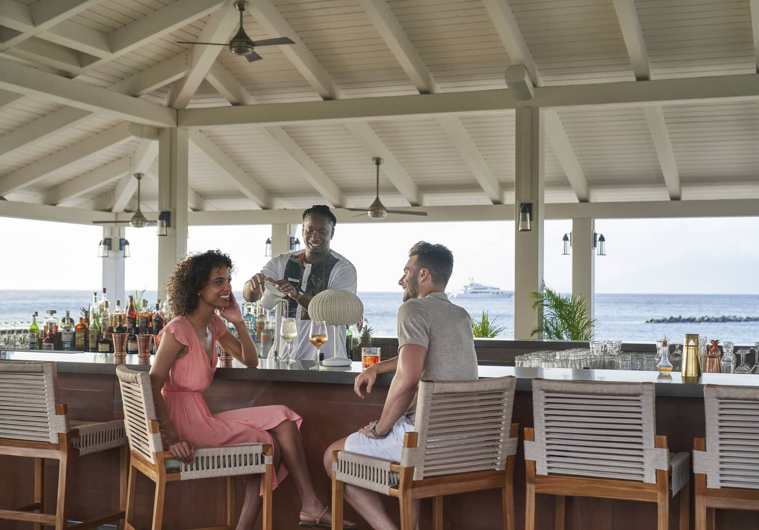 A man and woman talking at a bar outside while the bartender makes a drink.