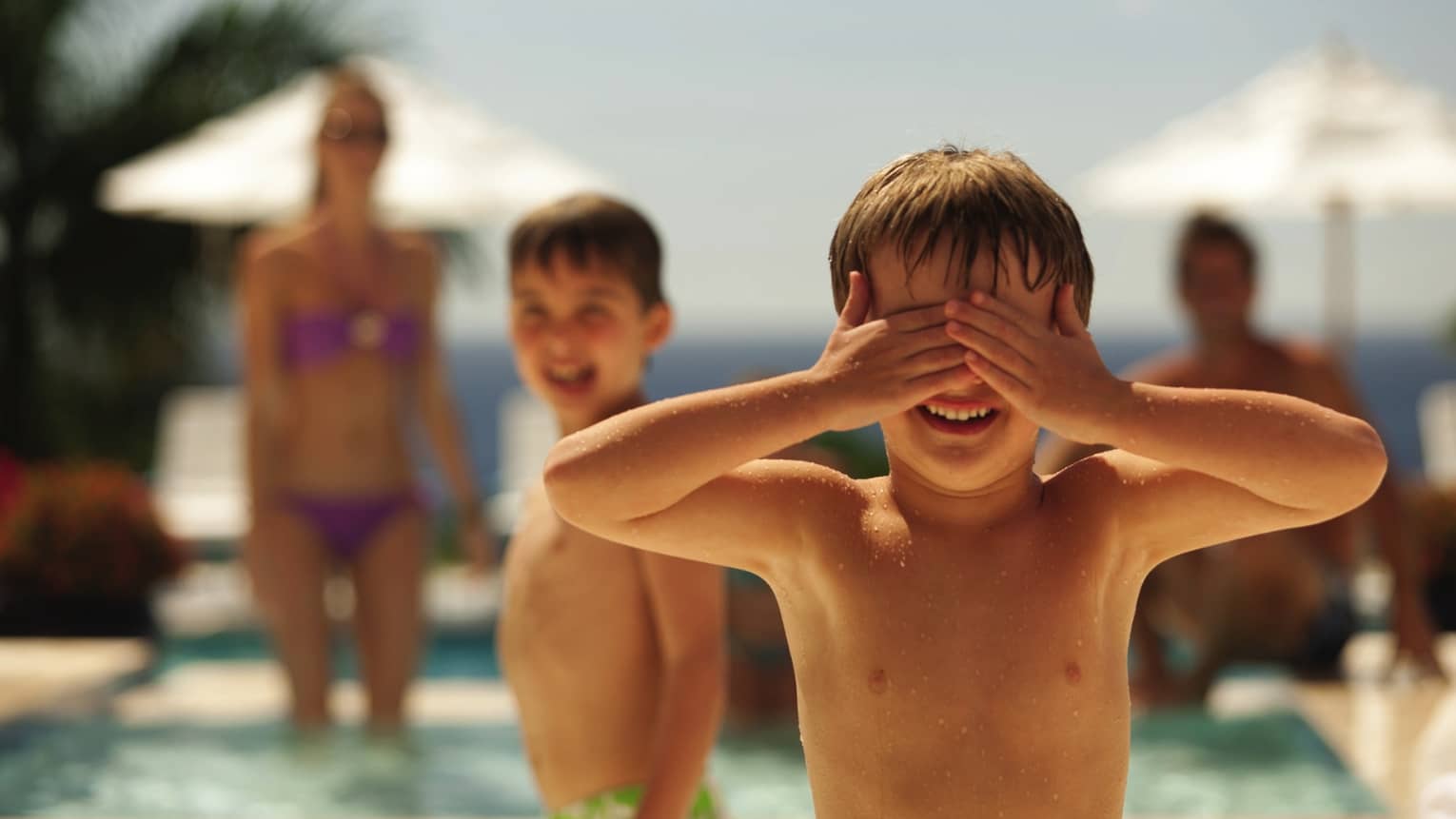 Young boy covers his eyes with his hands, family swimming in background
