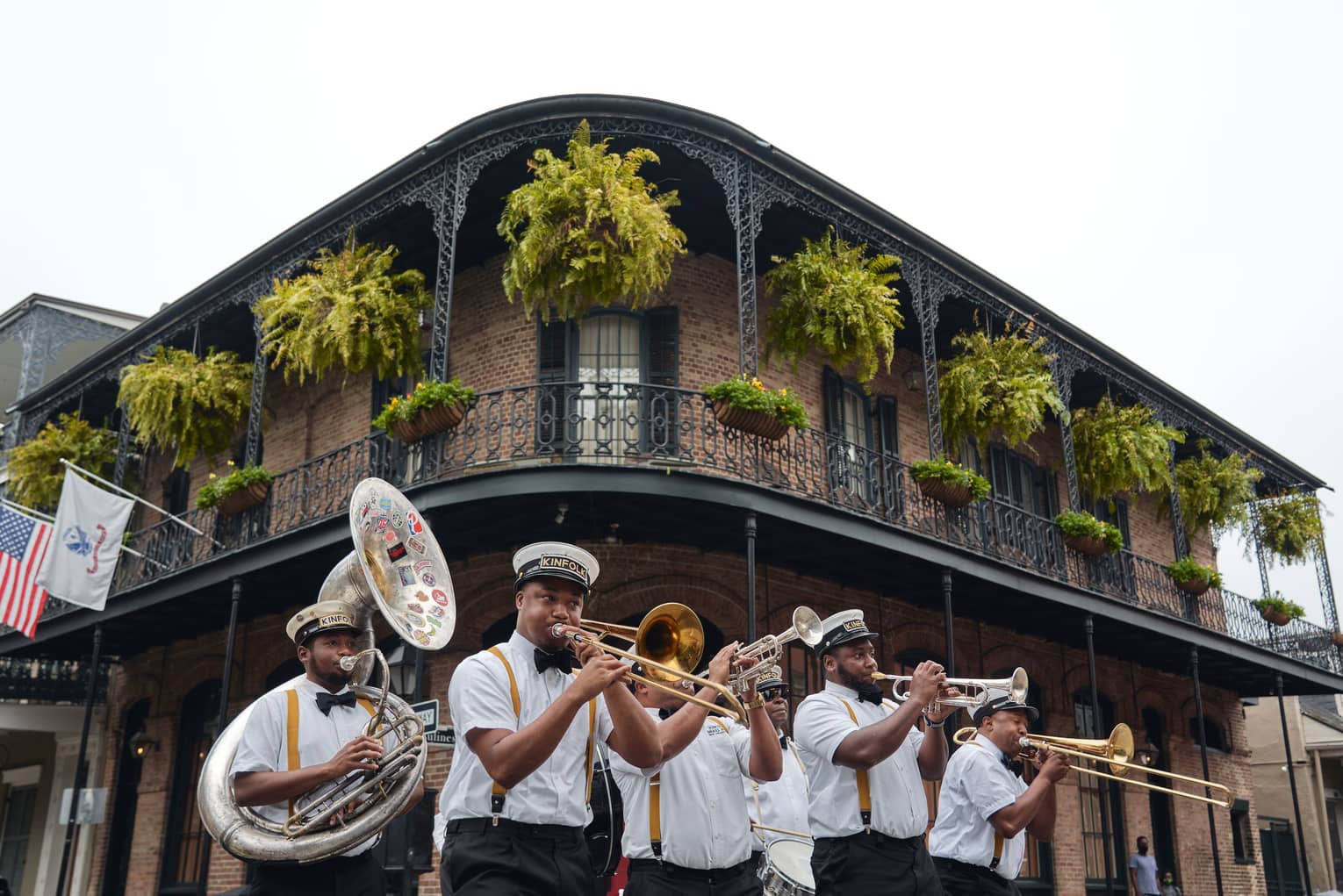 Six-piece brass band plays at the corner of a two-storey building beneath a balcony adorned with flags, ferns and flowers