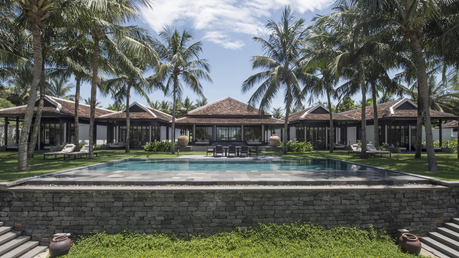 Luxurious villa with a large swimming pool, surrounded by palm trees and greenery, featuring modern architecture with tiled roofs and outdoor seating areas.