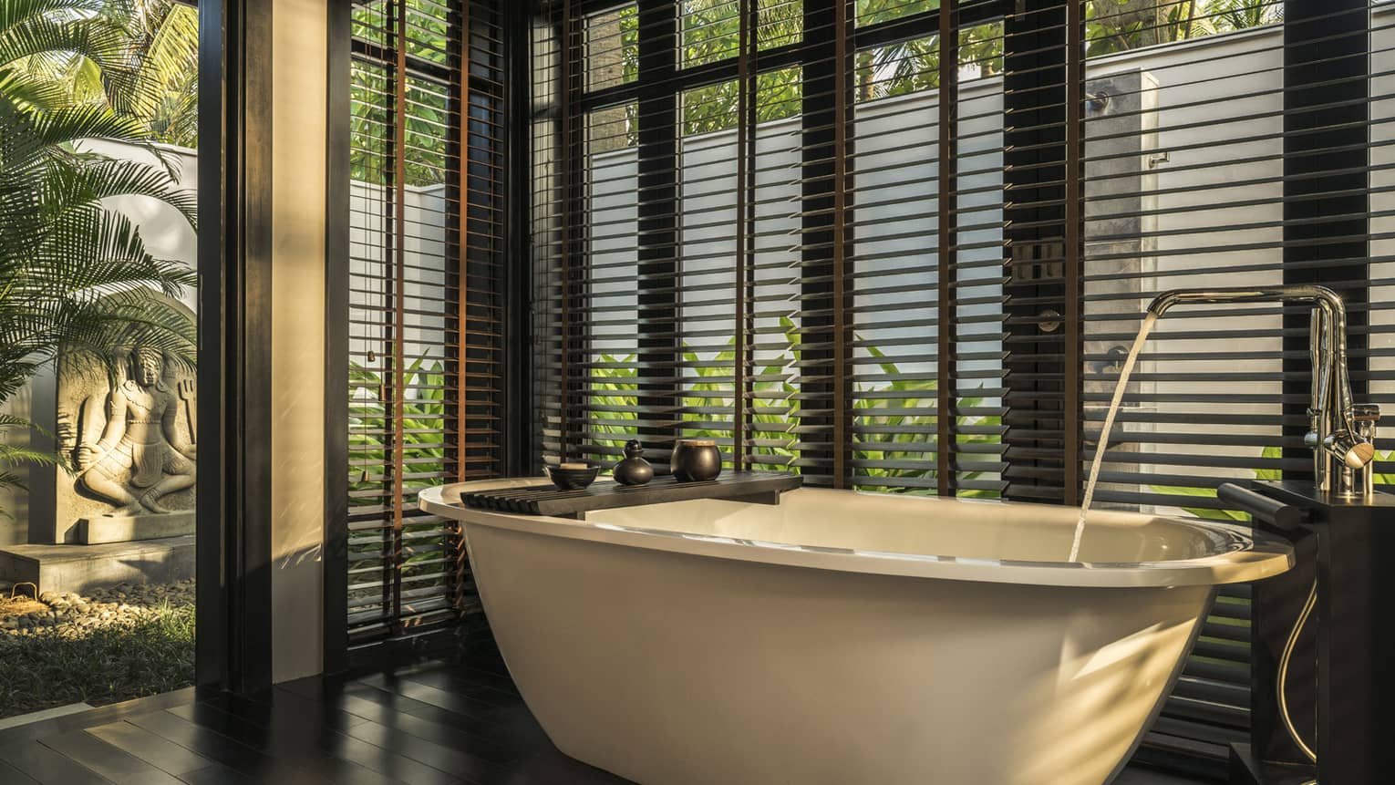 Luxurious bathroom with a free-standing bathtub, surrounded by large windows with wooden blinds, overlooking a garden with a stone sculpture and tropical plants.