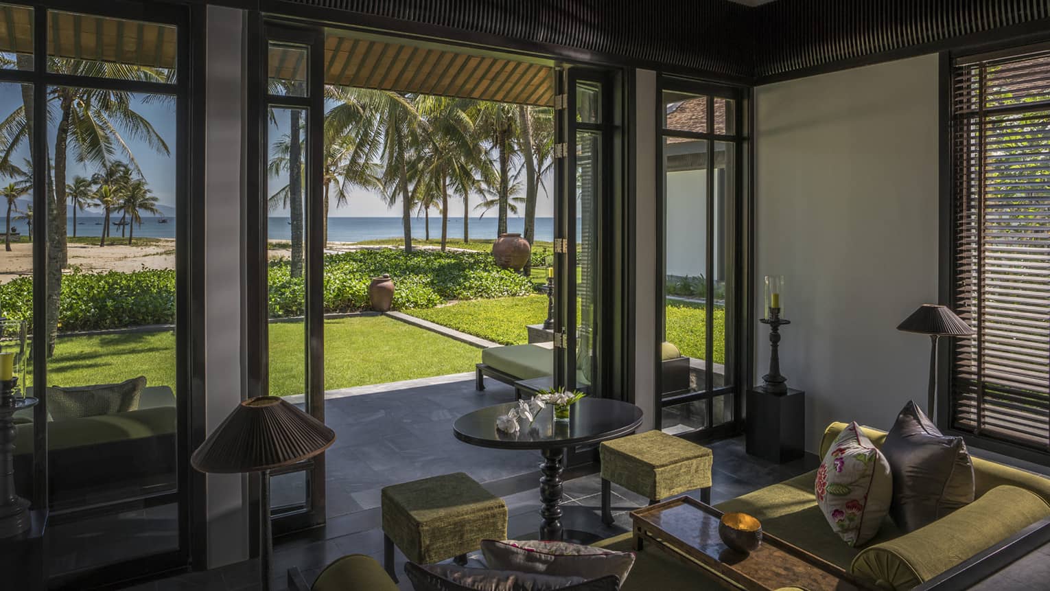 Luxurious villa living room with open doors leading to a private garden, overlooking a beach with palm trees and ocean views.