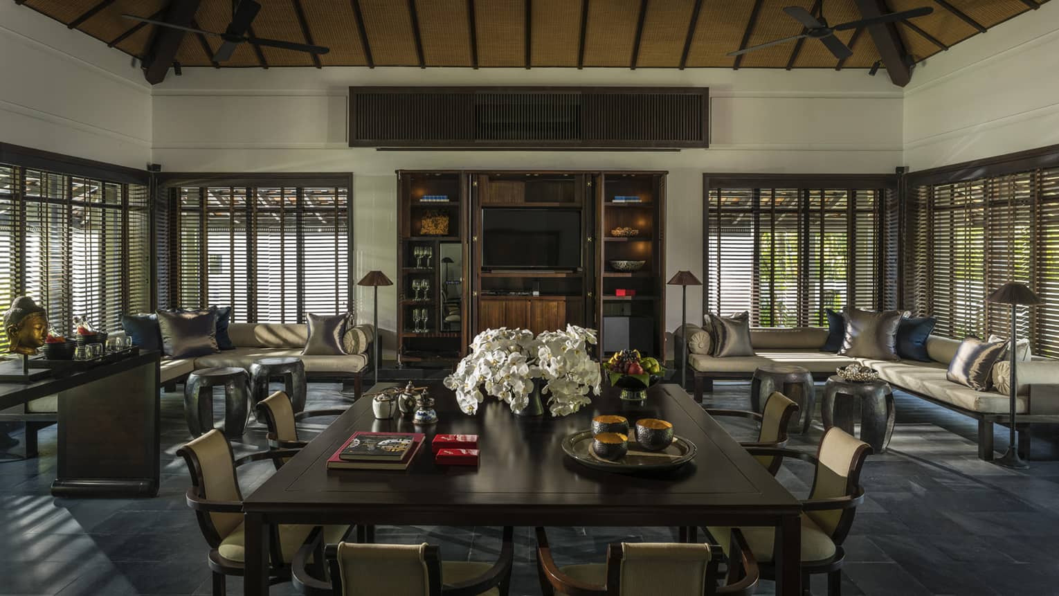 Large dining table with flowers in middle of room lined with sofas, windows, shutters