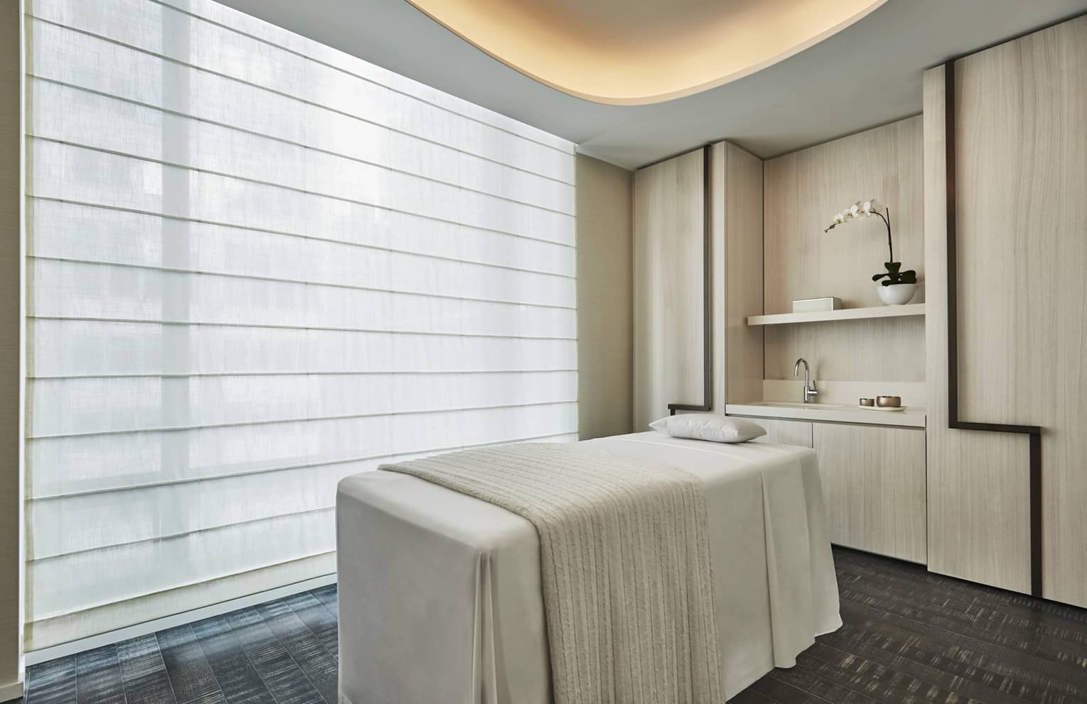 Spa massage table with white blanket by large window with shades, under light