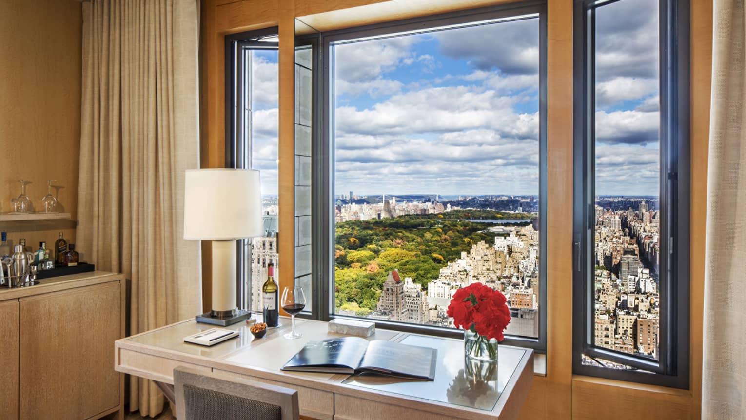 Writing desk with open book, glass of red wine and flowers under open window overlooking city, park