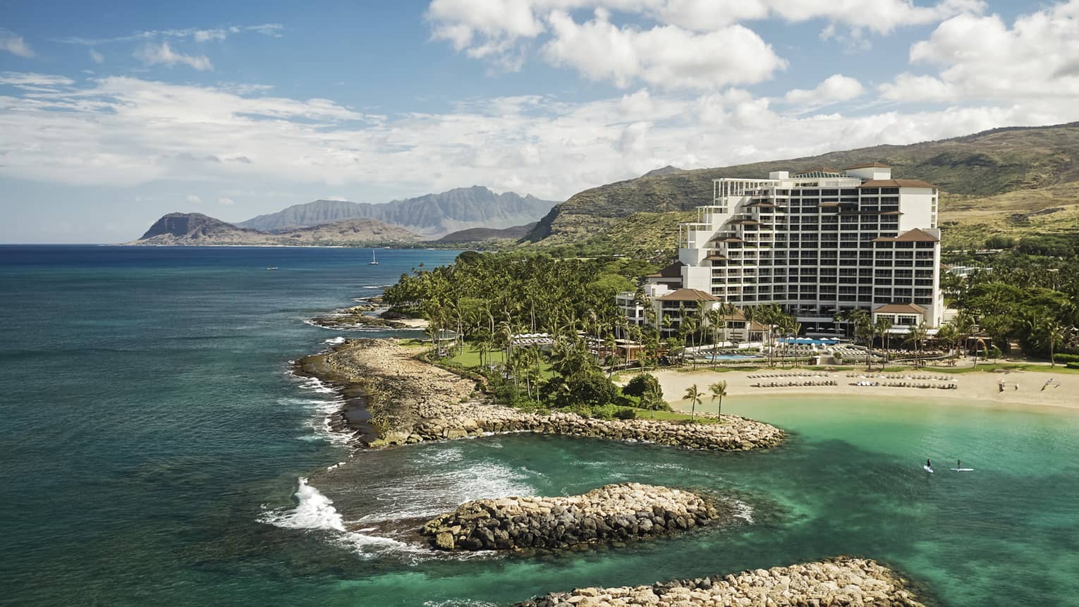 Tall white Four Seasons Resort Oahu building on mountainside by sand beach, rocks, ocean