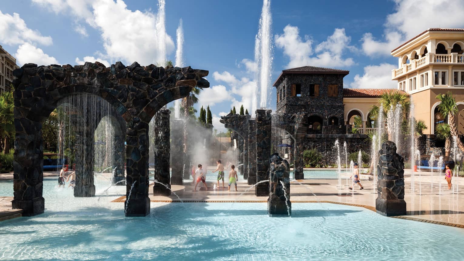 Kids play in water, under sprinklers and stone pillars in the Splash Zone water park