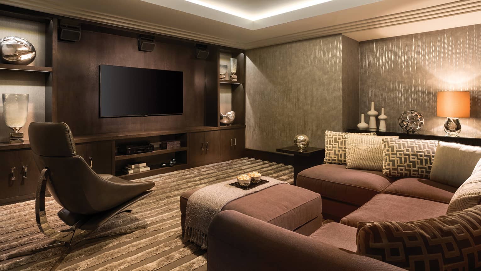 TV on wood panel wall. cabinet, across from L-shaped brown sofa, plush ottoman, modern leather chair