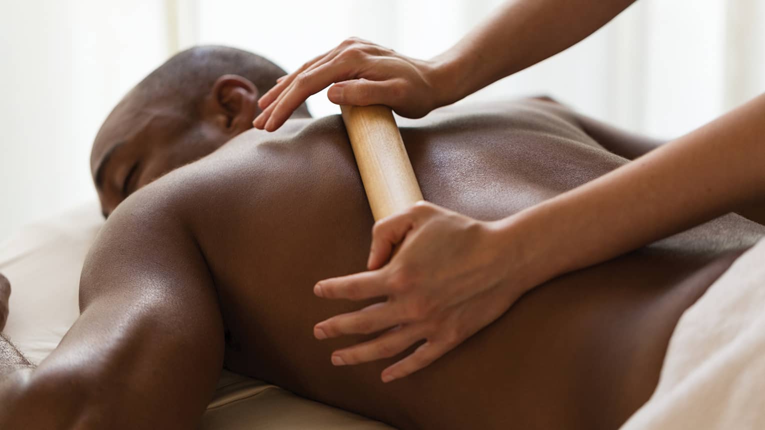 Masseuse rubs large heated wood baton across man's bare shoulders