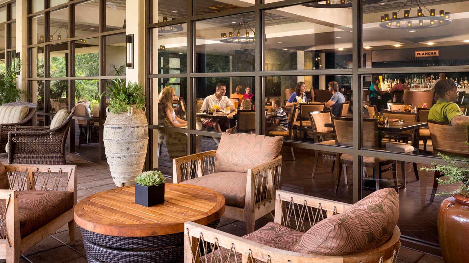 Plush patio chairs around wood table, tall vase, glass wall showing diners at tables