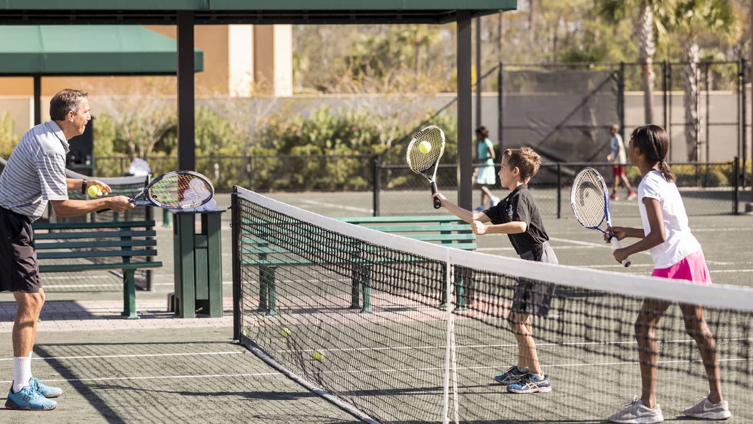 Man serves tennis ball to two kids on other side of net on small tennis court