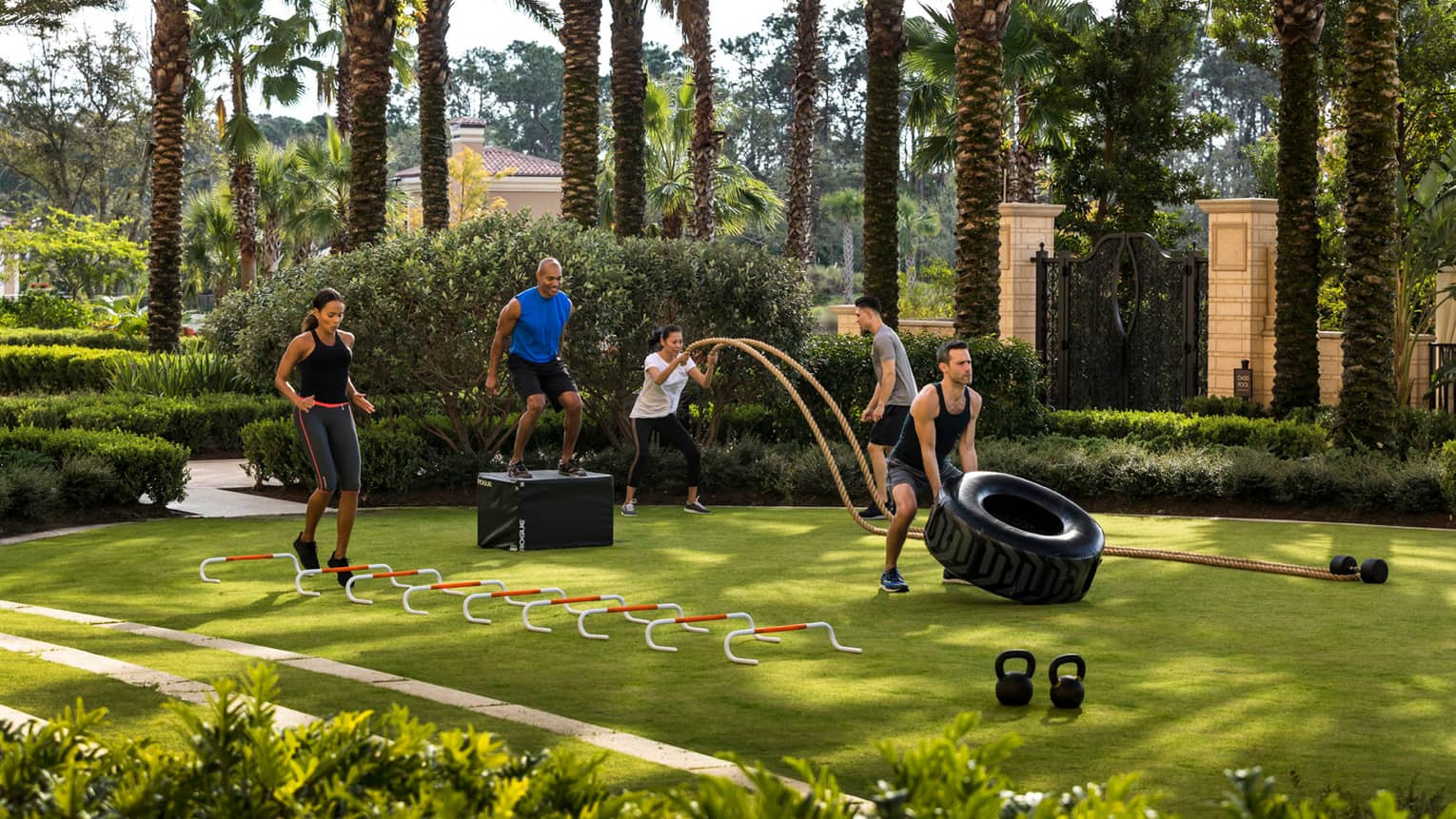 High intensity circuit training fitness course on green lawn, man lifts tire, woman jumps over hoops